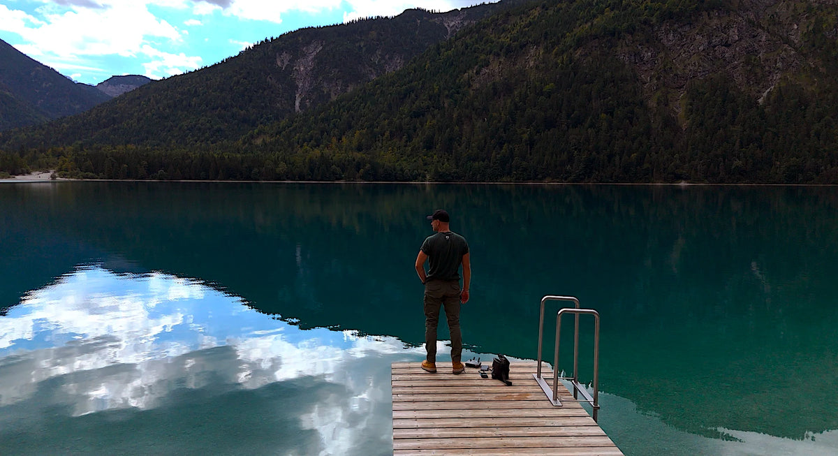 Sebastian Schneider steht auf einem Steg an einem Bergsee, Wolken spiegeln sich im Wasser