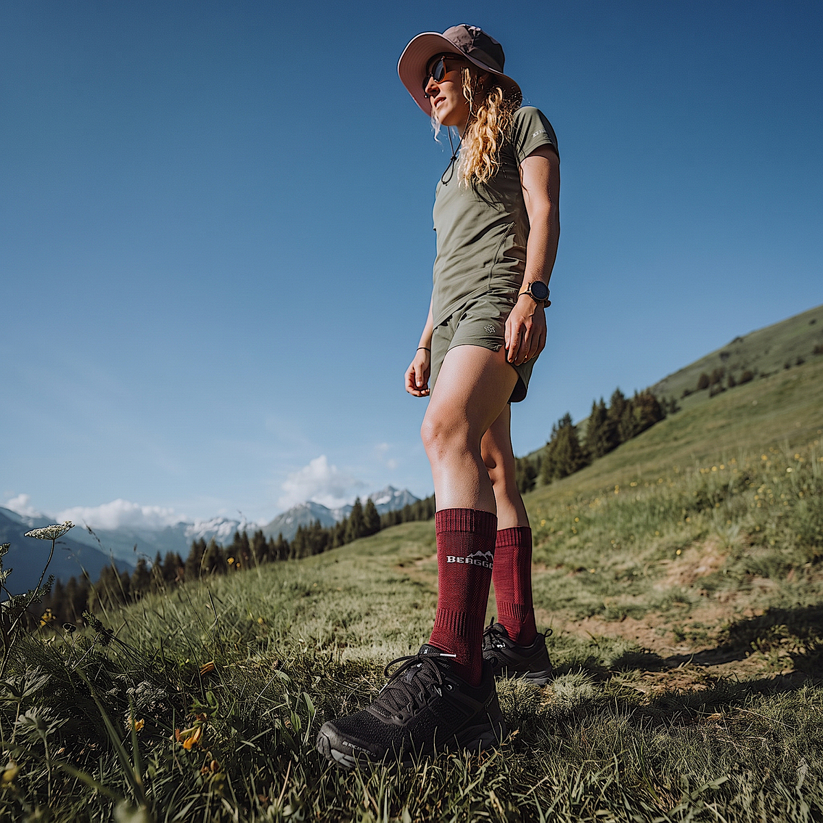Wanderin in Shorts und Sonnenhut steht am Berghang, trägt weinrote Merino Wandersocken und schwarze Trailschuhe
