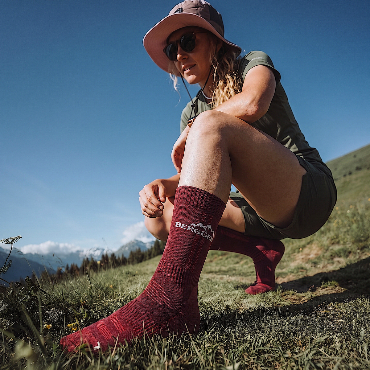 Frau hockt auf einer Almwiese in weinroten BERGGEIST Merino Wandersocken ohne Schuhe, blauer Himmel
