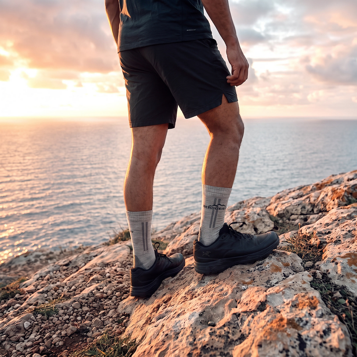 Wanderer in Shorts und schwarzen Schuhen mit hellgrauen Merino Wandersocken auf Felsklippe am Meer