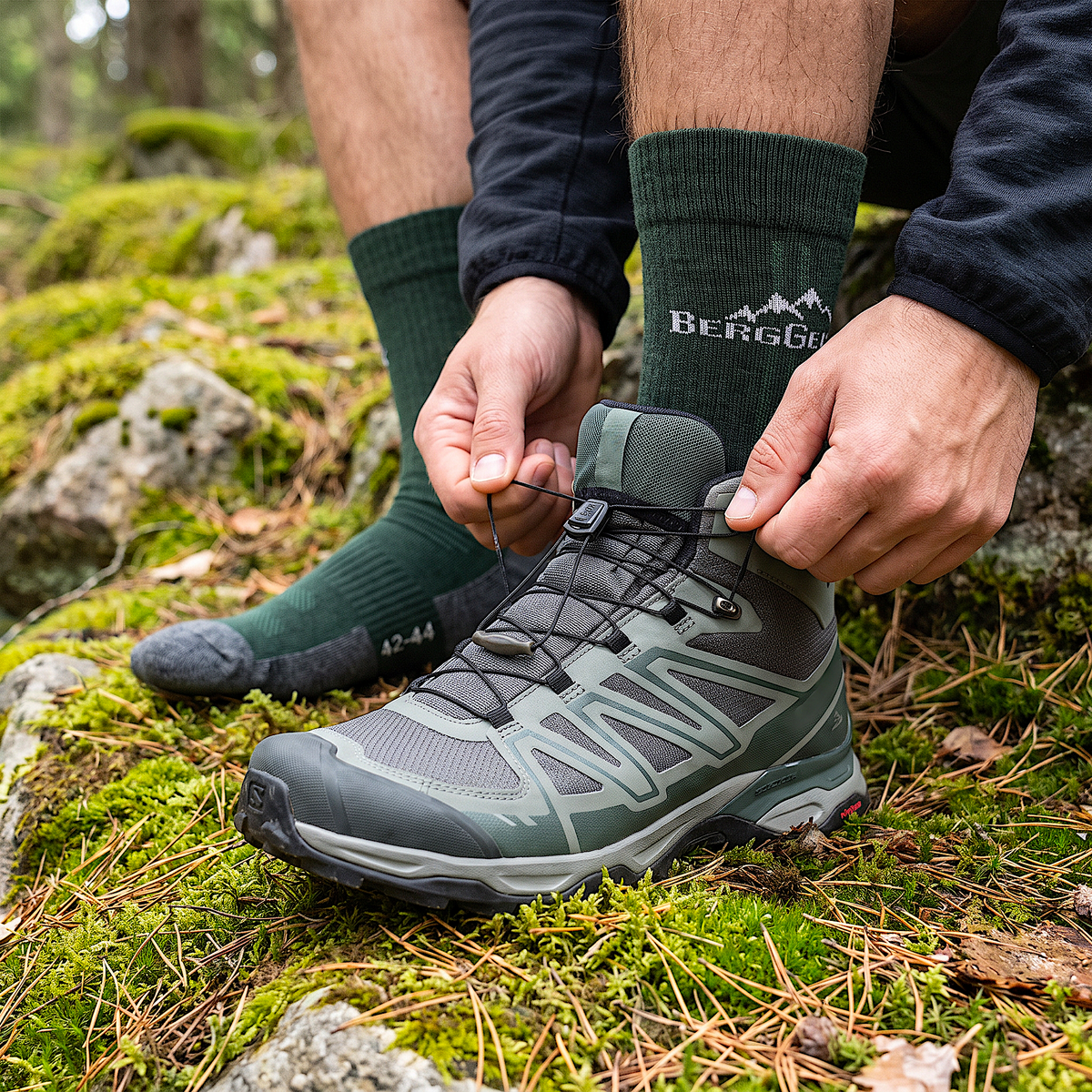 Hände schnüren grauen Wanderschuh, dunkelgrüne Merino Wandersocken mit grauer Spitze auf Moos und Kiefernnadeln