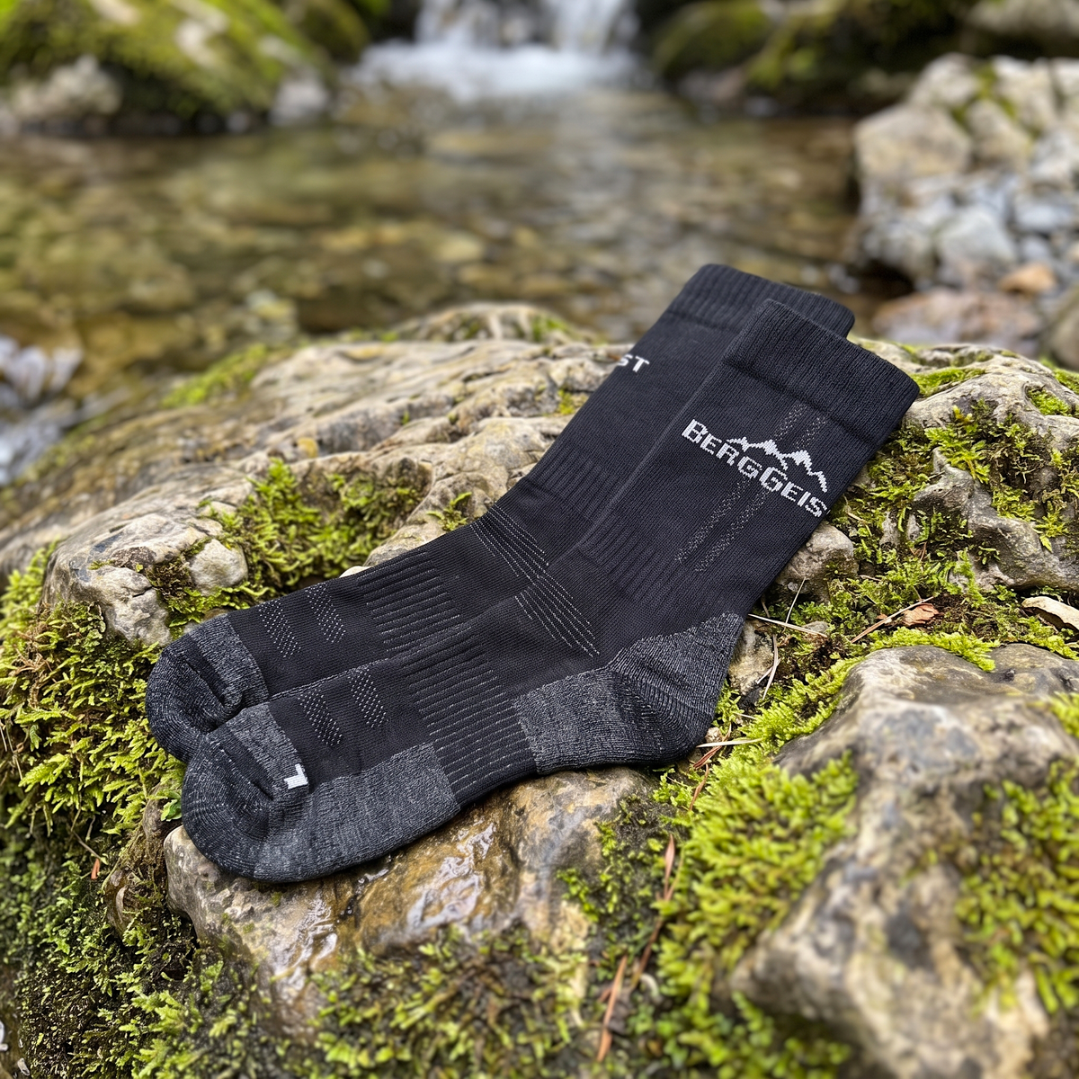 Paar schwarze Merino Wandersocken mit BERGGEIST-Logo auf moosbewachsenem Stein an einem Bach